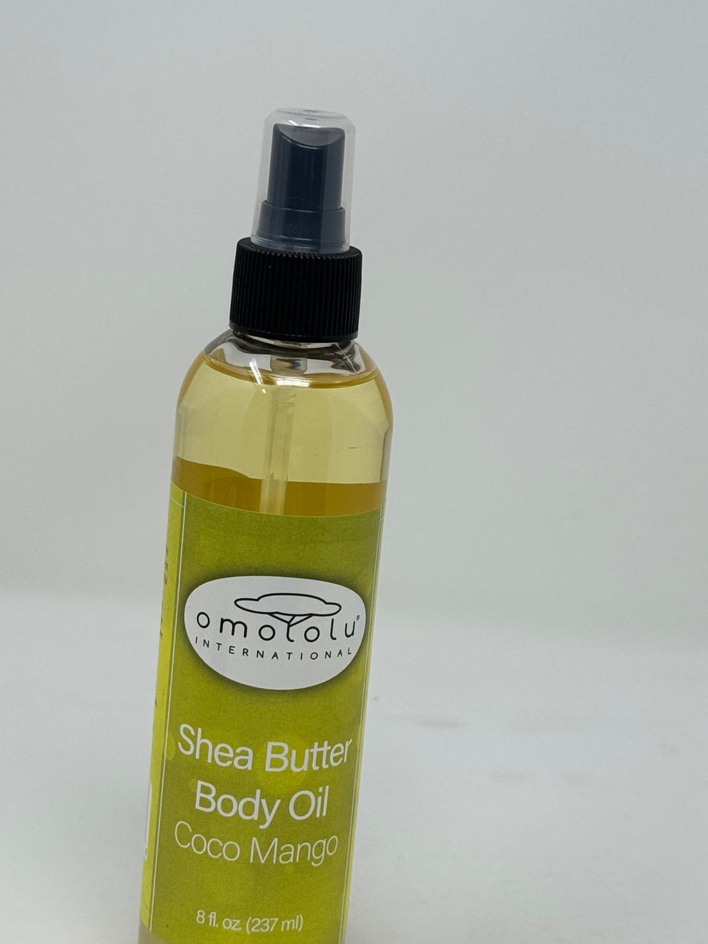 A bottle of Omotolu International Shea Butter Body Oil in Coco Mango scent, featuring a yellow-green label and a black spray nozzle, set against a plain light gray background.